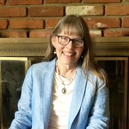 Lynn Perez-Hewitt head shot. Woman smiling wearing glasses with long hair. She is wearing a blue blazer over a white shirt.