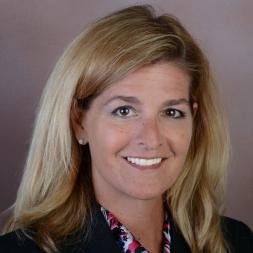 Natalie Glumm headshot. Woman smiling with blonde hair. She is wearing a black blazer with a pink and white scarf.