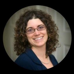 A woman wearing glasses, curly brown shoulder length hair, and a blue blazer.