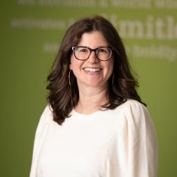 A woman wearing glasses, shoulder length brown hair, and a white long sleeve blouse. She is standing in front of a green background with blurred white lettering.