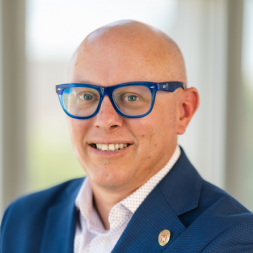 A man wearing glasses and blue suit jacket with a white collar shirt under.