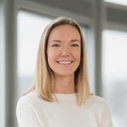 Courtney Spilker Headshot. A Woman with blonde shoulder length hair smiling at the camera. She is wearing a white sweater.