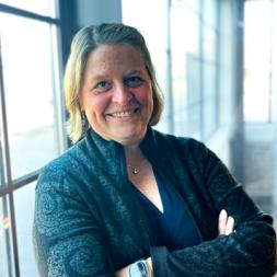 Dani Luckett photo. A woman is smiling with shoulder length blond hair. She is wearing a black jacket and has her arms folded across her chest.