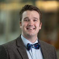 Pete Schweiger headshot. A man wearing a brown suit jacket, blue collar shirt, and a blue bow tie. The background is blured.