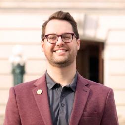 A man smiling, wearing glasses. He is wearing a maroon blazer with a gray collared shirt. The man has short brown hair and glasses.