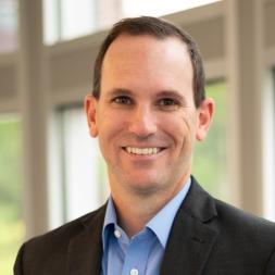 A man standing in front of a window. He has short brown hair, wearing a blue collared dress shirt and a blue business suit jacket.