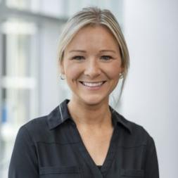 A woman smiling wearing a black collared shirt. She has blonde hair pulled back in a ponytail.