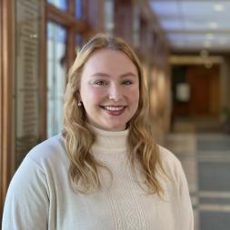 A woman smiling with long blonde hair. She is wearing a white turtleneck sweater and standing in front of a long hallway.