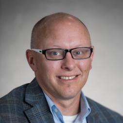 A man wearing glasses and blue suit jacket with a blue collar shirt under.