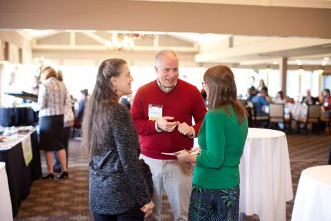 Three people talking at a luncheon event. Two women have their backs to us and a man is in the middle facing the camera.