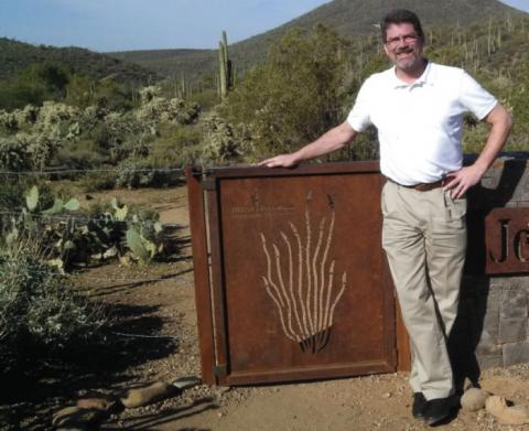 Man with a white shirt and khaki pants standing near a metal gate with a desert behind him.