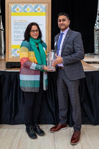 Two people (a woman on the left and a man on the right) stand in front of a stage. They are both holding an award.