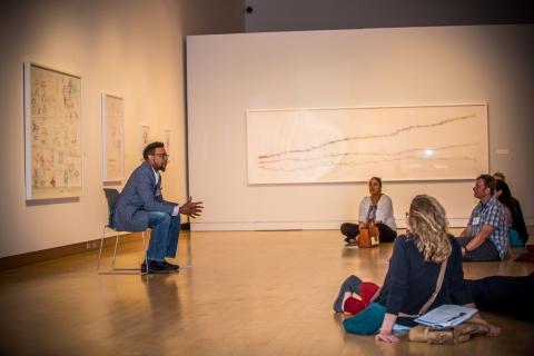 A man sits in a chair talking with a small group of individuals sitting on the floor. The background is white walls with artwork.