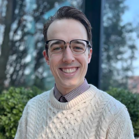 A man standing in front of trees. He is smiling with short brown hair. He is wearing glasses and a white sweater.