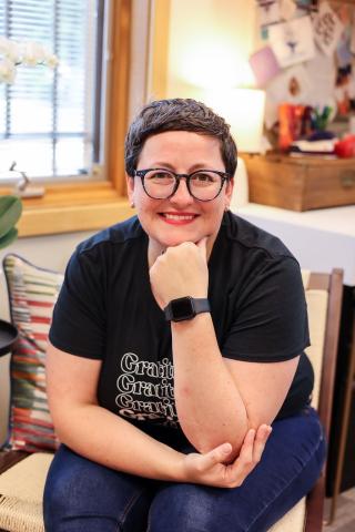A woman is sitting down with her face resting on her hand. She is wearing a black shirt, brown glasses, and has short brown hair.