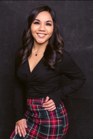 Jamie Perez photo- a woman smiling and posing for the camera. She is wearing a black shirt and plaid skirt with her hand on her hip. There is a black background.