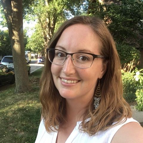 Woman smiling in front of a background with grass, trees and a street with parked cars. Sarah is smiling, she has red hair, black-rimmed glasses, and is wearing a white sleeveless shirt. 