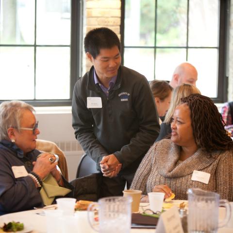 Two women sitting and talking at a table. A man is leaning down to speak with them.