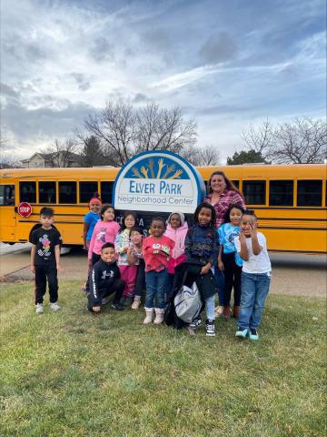 A group of children standing around a sign that reads "Elver Park Neighborhood Center". There is a school bus in the background.