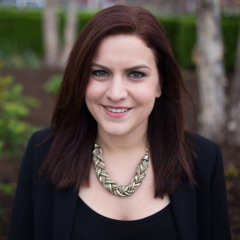 Woman smiling in front of a blurred nature background. The woman has dark hair and she is wearing a black shirt, black blazer, and a gold chain necklace.