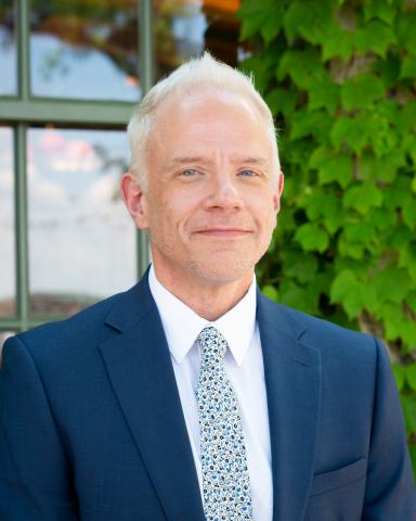 A headshot of a man. He is wearing a blue suit coat over a white shirt with a blue speckled tie.