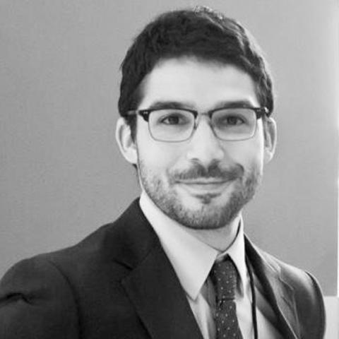 Black and white photo of a man wearing a suit and smiling. He has short black hair, a short beard and is wearing glasses.