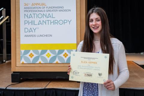 Eliza, a young girl with long brown hair and smiling, stands in front of a podium holding her award sign.