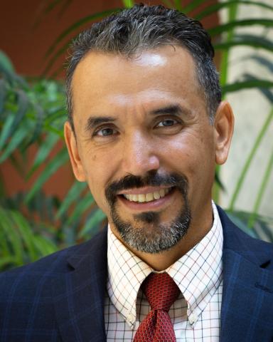 Headshot of a man wearing a blue suit and red tie. The background is a green palm plant.