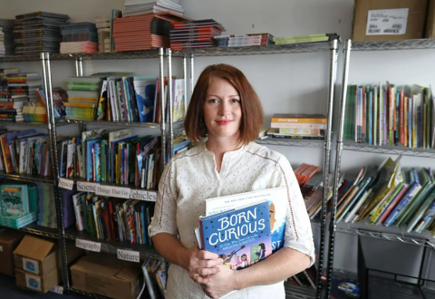 A woman stands smiling. She is holding books in her hands and racks of books are behind her.