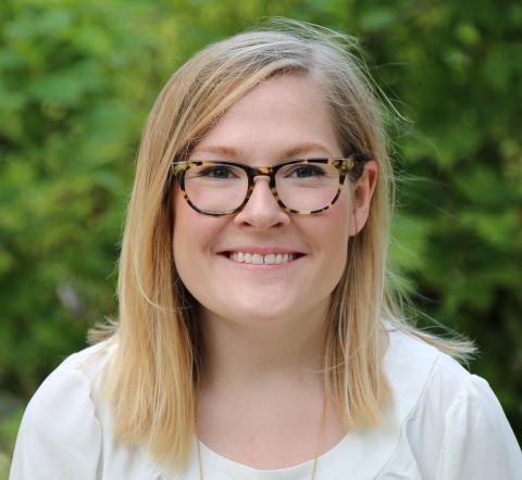 A photo of a woman with short blonde hair and wearing a white blouse. She is wearing glasses.