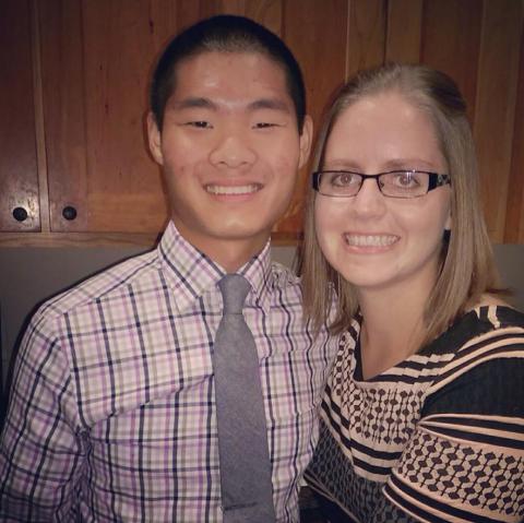 A man and a woman standing close to each other and both smiling. The man is on the left wearing a plaid shirt and tie. The woman is on the right wearing a black and beige shirt and has glasses.