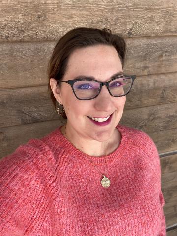 Woman smiling and leaning against a wood wall. She is wearing glasses and a red sweater.