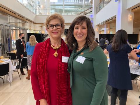 Image of two woman posing among a backdrop of people talking. The woman on the left is wearing glasses and a red long sleeve dress. The woman on the right has shoulder-length brown hair and wearing a long-sleeve green dress.