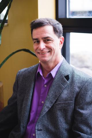 A man smiling for a headshot photo. The man has short dark hair and is wearing a gray suit jacket with a purple dress shirt.