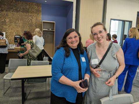 Two women are standing in front of tables and people are mingling in the background. The woman on the left is wearing a blue sweater over a black dress. She has long dark hair. The woman on the right is wearing a grey and white striped dress. She is wearing her hair pulled back and glasses.