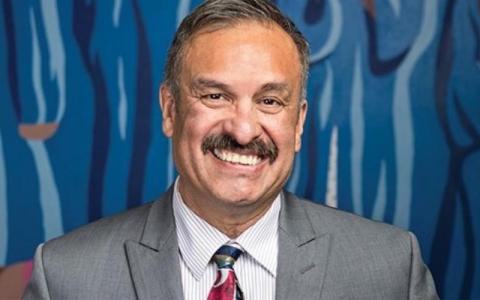 A headshot of a man smiling. He is wearing a grey suit jacket with a white shirt and tie. There is a blue background with streaks of varying shades of blue.