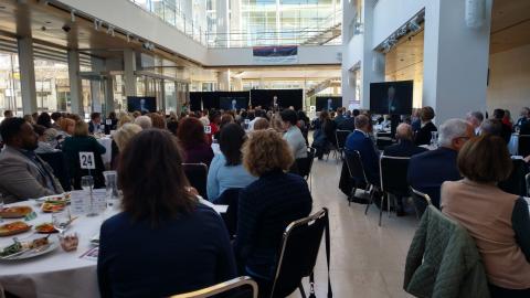 People sitting at tables in a long bright room. People are looking forward toward a stage.