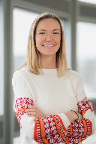 A woman standing with her arms folder. She has shoulder length blonde hair and is wearing a white sweater with orange detail on the wrists.