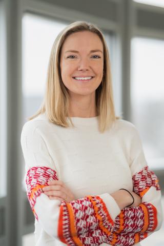A woman standing with her arms folder. She has shoulder length blonde hair and is wearing a white sweater with orange detail on the wrists.