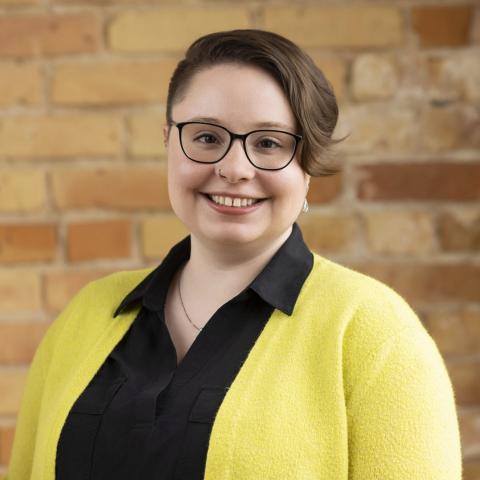 A woman is standing in front of a brick wall. She has ear length short brown hair. She is wearing black rimmed glasses, a yellow sweater over a black collared shirt.