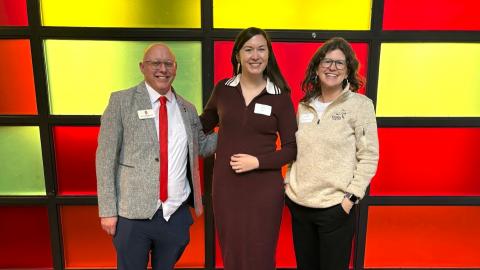 Three people standing in front of a yellow and red wall. There is a man on the left with a women in the center and another on the right. 