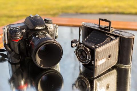 Two cameras sitting on a reflective surface with a green area blurred in the background.