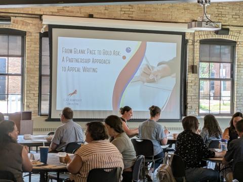A group of people sitting at round tables. A screen in the background reads "From blank page to bold ask: A partnership approach to appeal writing."