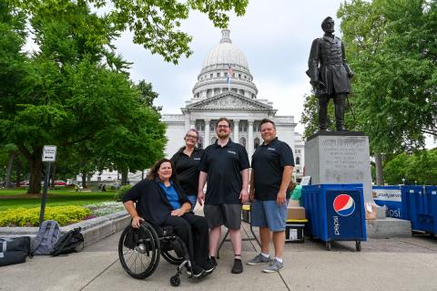 A group of four individuals in front of the Wisconsin state capitol building. One woman is in a wheelchair and the other three individuals are standing next to her.