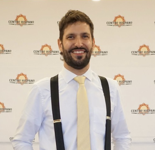 Man standing and smiling. He is wearing a white shirt, yellow tie, black suspenders, and gray slacks.