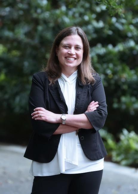 A woman in standing in front of a background with trees. She is crossing her arm and wearing a black blazer over a white shirt.