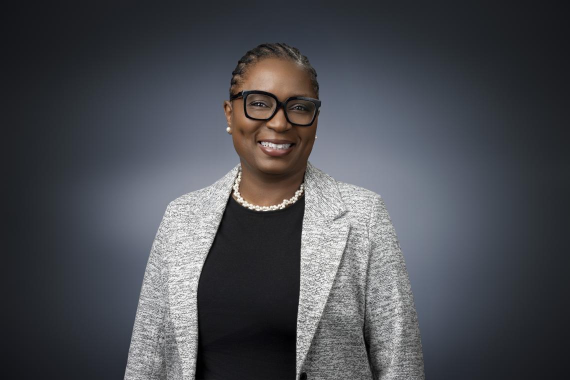 A woman is posed in front of a black and gray backdrop. She is wearing a gray blazer, black shirt, and black glasses.