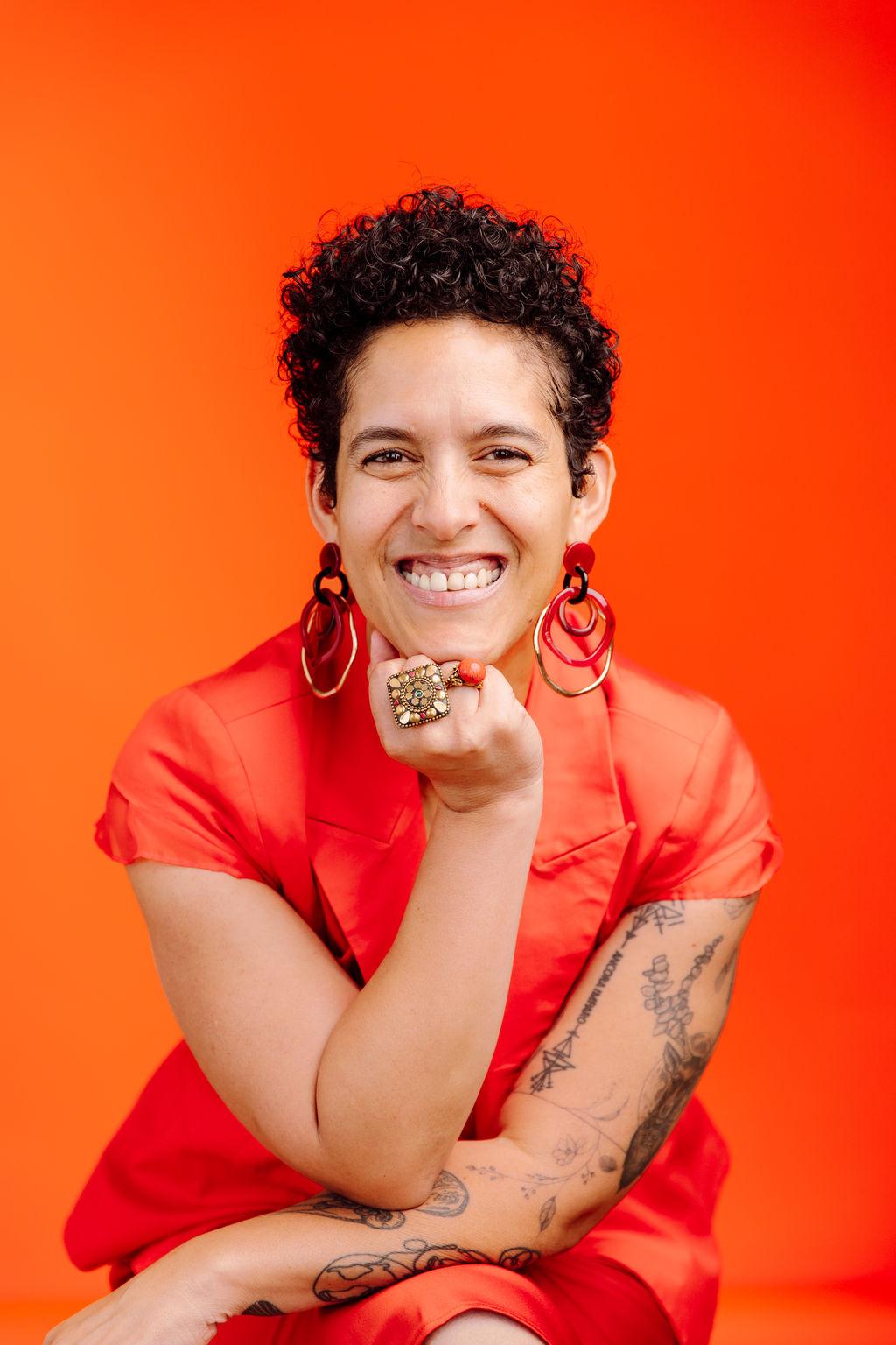 An orange background with a woman sitting with her hand on her chin. She is wearing a red dress.