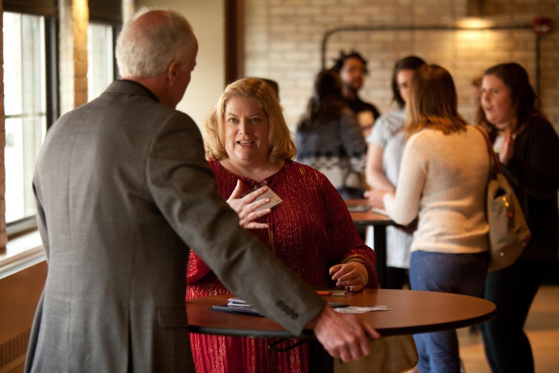 Man wearing a suit with his back to us. Speaking to a woman in a red shirt with short blonde hair.